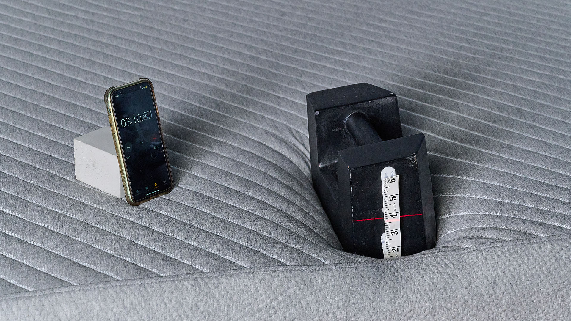 A close up of the 25kg weight balanced on the foot of the Siena Mattress for an edge support test, taking place in our sleep studio. There's a tap measure on the end of the weight and a red line, generated by a laser out of shot, indicates that the weight has sank roughly 3.6 inches into the mattress. To the left, a phone with a stopwatch indicates the test has been going on for 3 minutes 10 seconds.