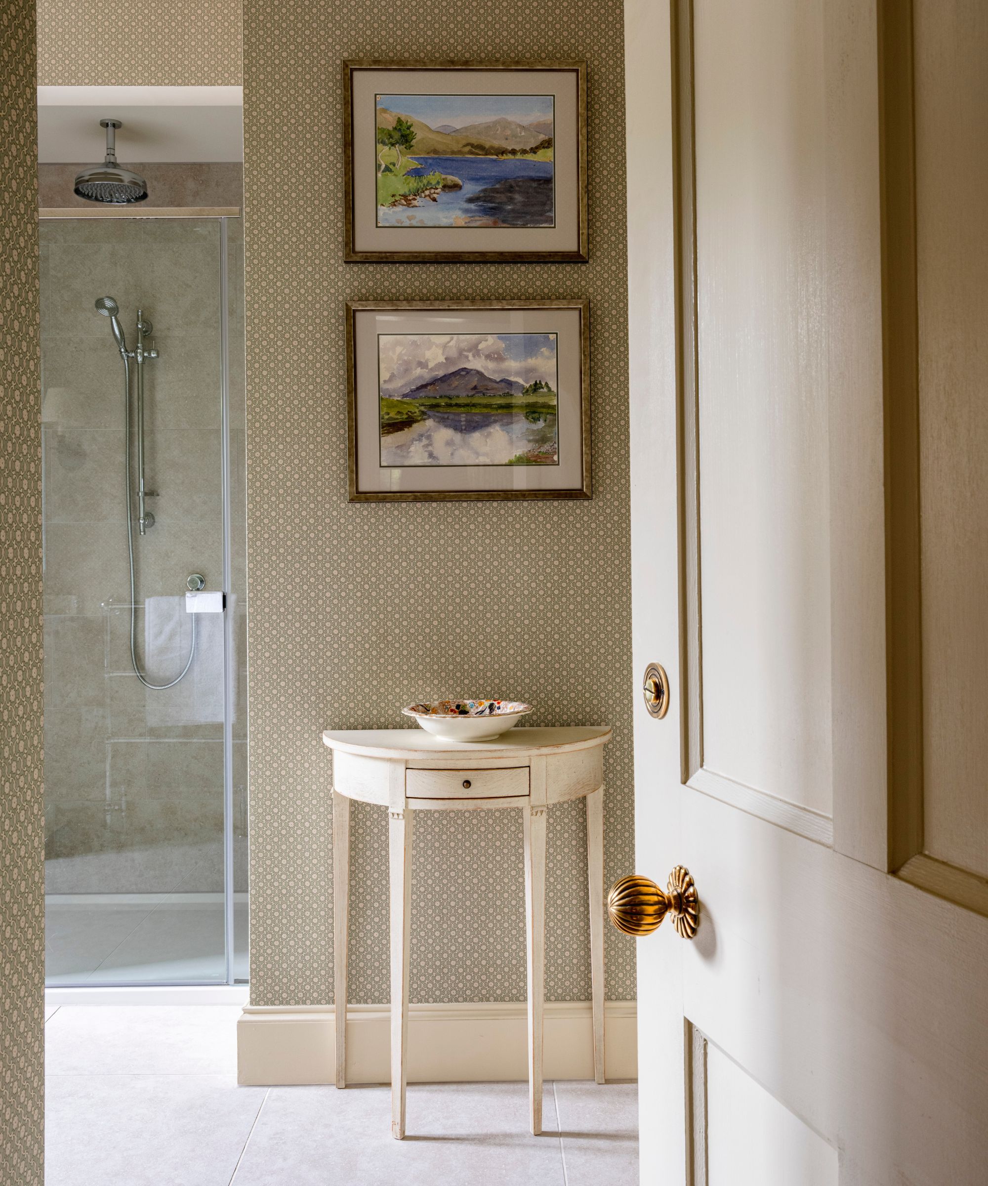 Vintage demi-lune console table with a single drawer styled beneath two framed landscape paintings, set against patterned wallpaper beside a glass-enclosed shower.