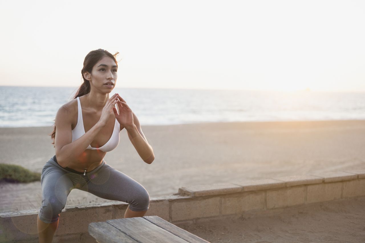 A woman performing a squat as part of a bodyweight workout by the beach 