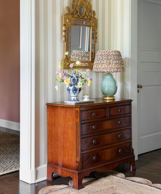 hallway with striped wallpaper and a scalloped jute rug with an antique mahogany sideboard styled with a blue table lamp and a vase of flowers