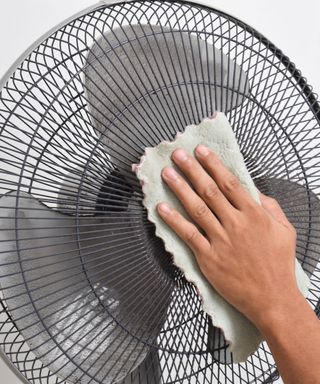 A close-up of a gray metal fan with a hand cleaning it with a mint green wavy edged cloth