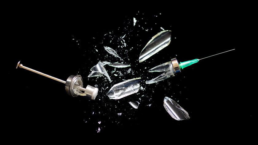 photo of a syringe for administering vaccines being smashed against a black backdrop