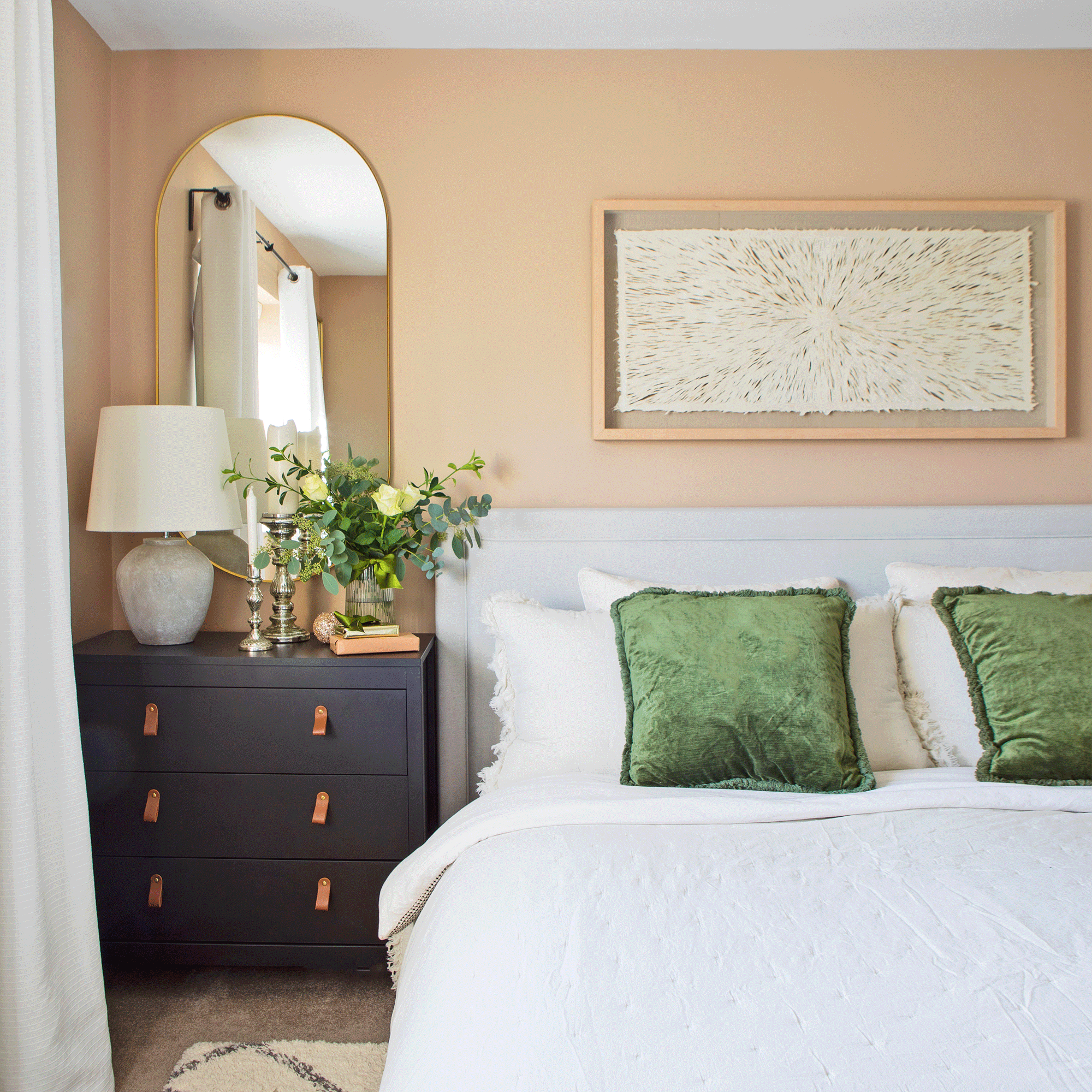 a pale peach painted bedroom with white bedding a green velvet cushions beside a black chest of drawers