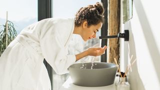 A woman stands in front of her sink wearing a bath robe in a brightly lit bathroom. There's water coming from the faucet which she holds in her cupped hands, ready to splash onto her face