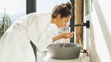 A woman stands in front of her sink wearing a bath robe in a brightly lit bathroom. There's water coming from the faucet which she holds in her cupped hands, ready to splash onto her face