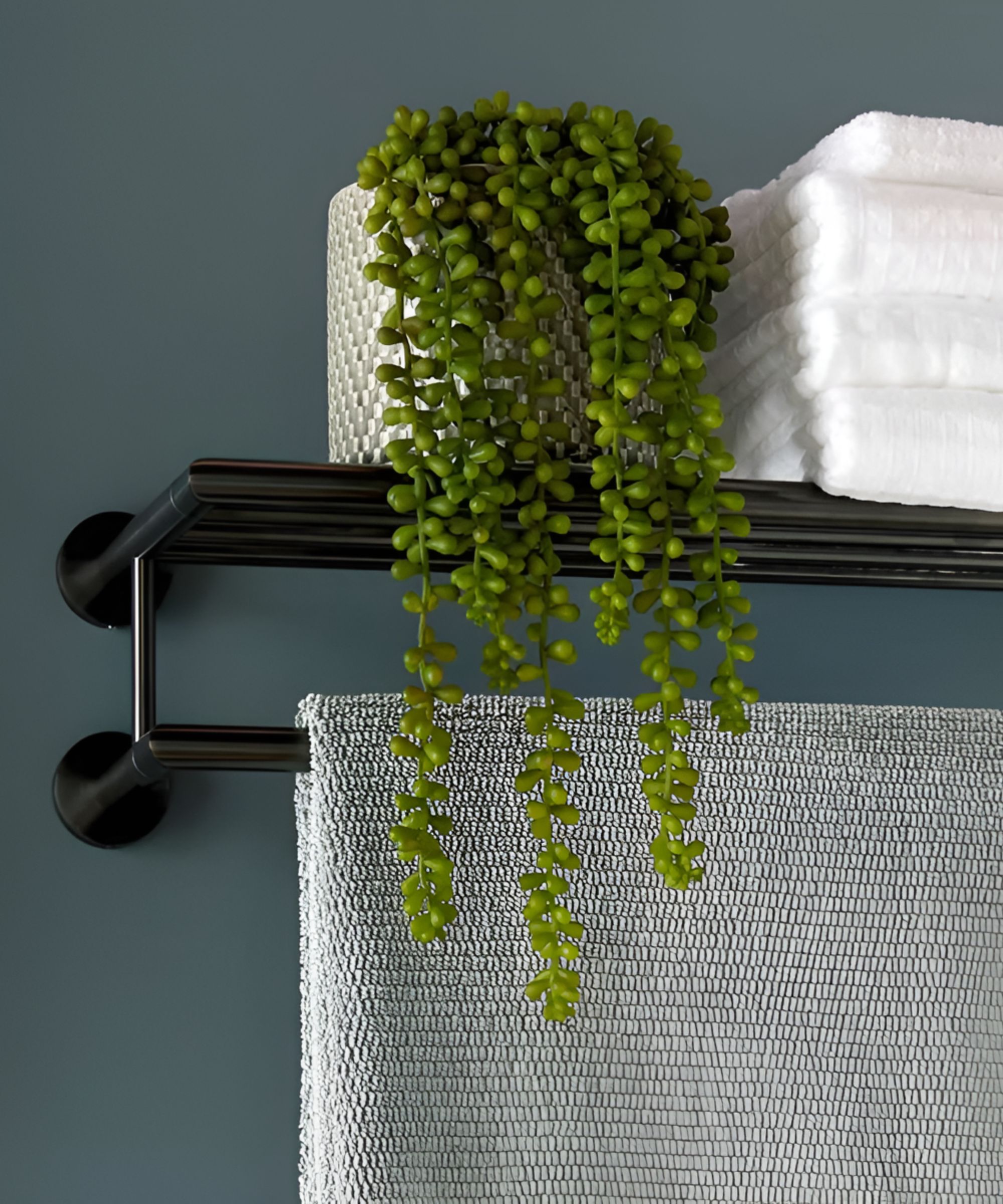 A black wall-mounted rail doubles as a slim shelf, styled with folded white towels and a trailing plant. The dark finish contrasts neatly with the deep blue wall.