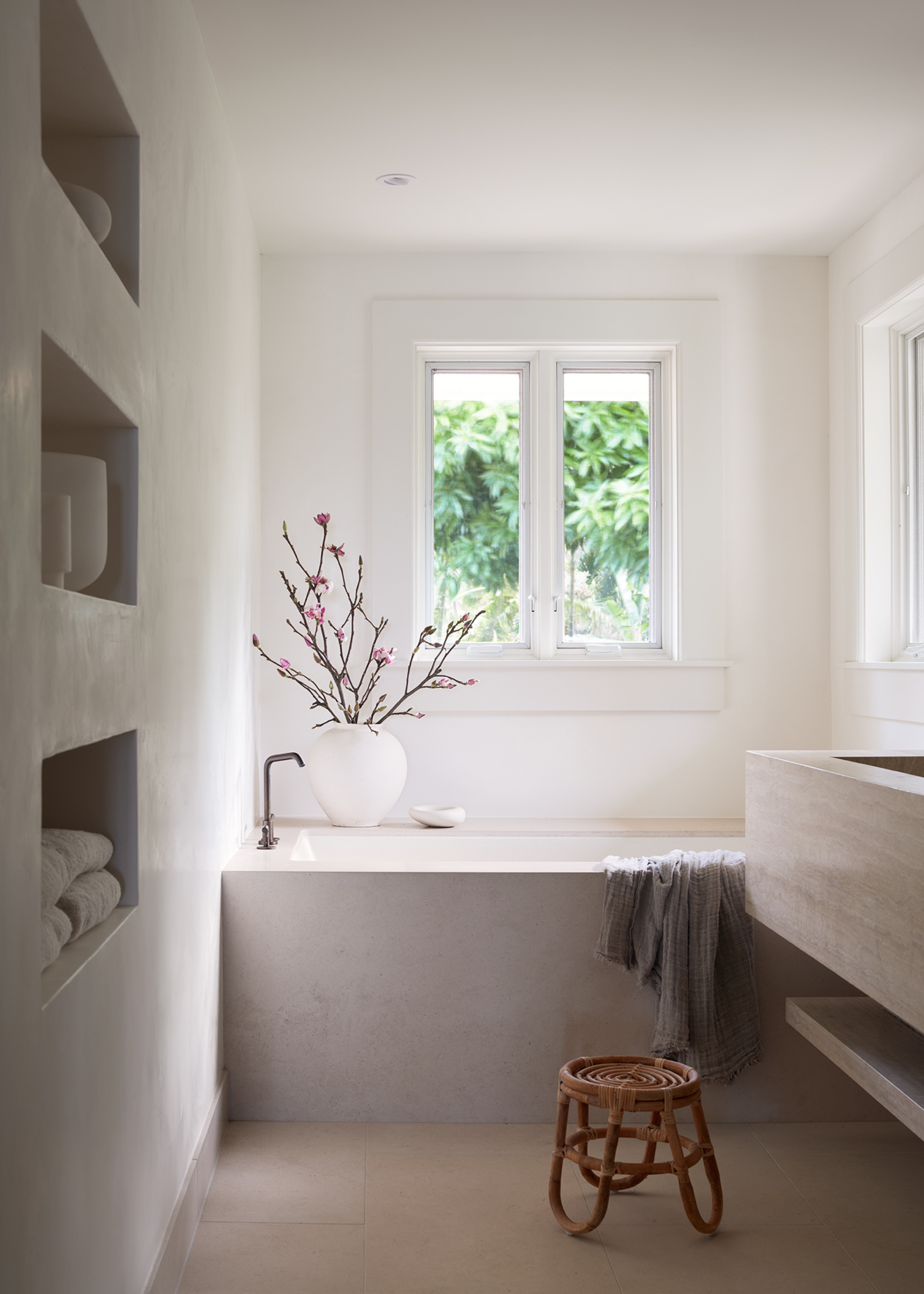 A white bathroom with in-built shelving nooks, a wicker stool, a stone basin and a vase of stems