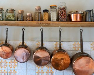 Copper pans hanging from a mounted wall shelf in a country kitchen a kitchen, going from smallest to largest, with shelf above filled with jars of different ingredients