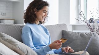 A woman sits on the sofa, credit card in hand, smiling as she uses her laptop