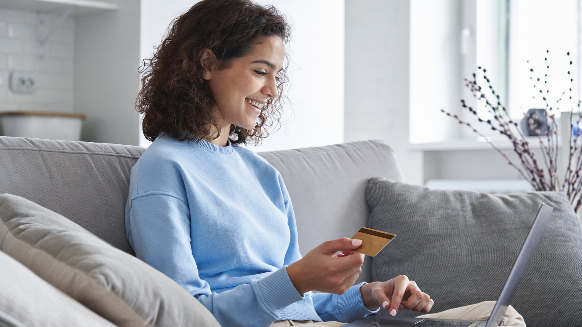 A woman sits on the sofa, credit card in hand, smiling as she uses her laptop