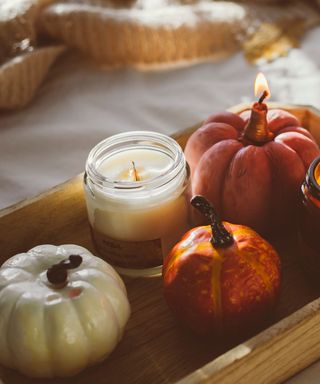 An unlit jar candle on a wooden tray next to a small pumkin and a lit pumpkin shaped candle