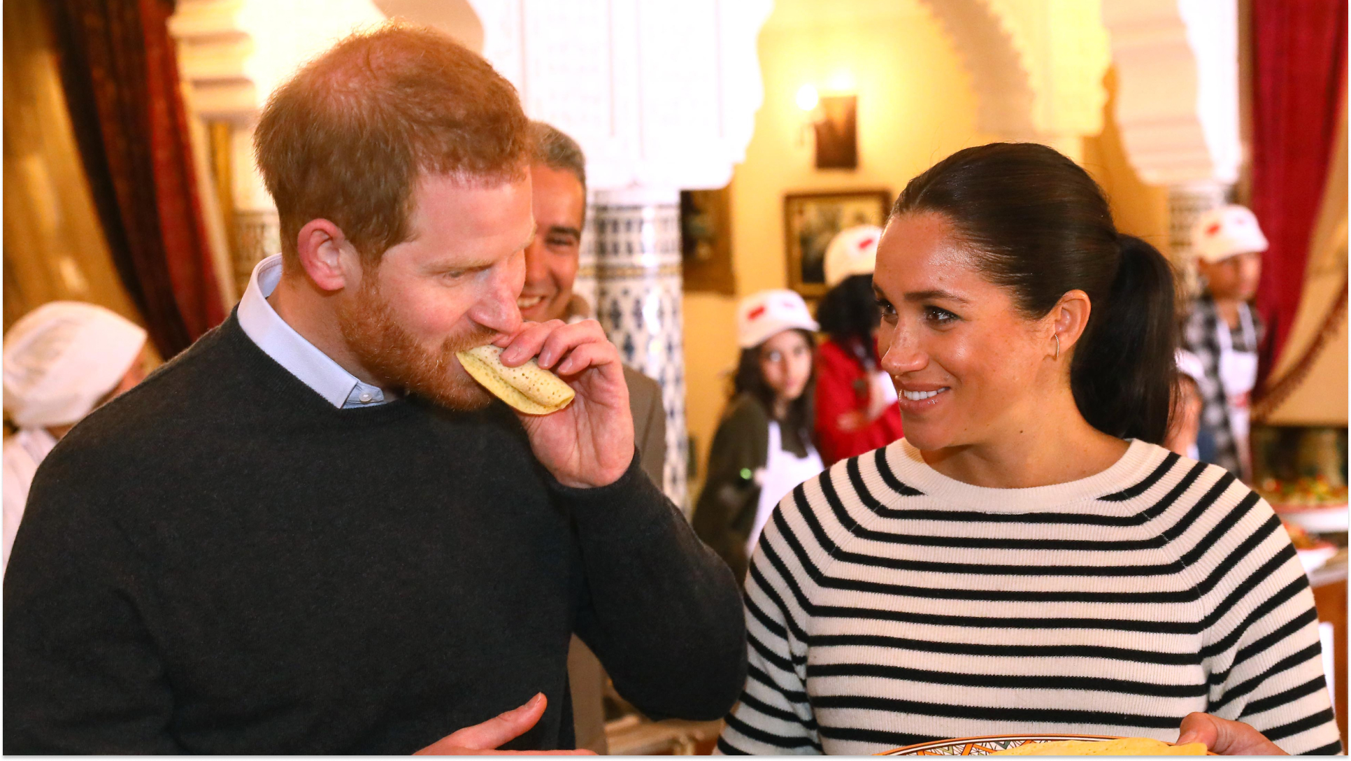Prince Harry eats a sandwich while Meghan Markle, wearing a striped top, watches