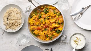 Aerial shot of cauliflower curry in a saucepan next to a bowl of rice and smaller bowl of natural yogurt