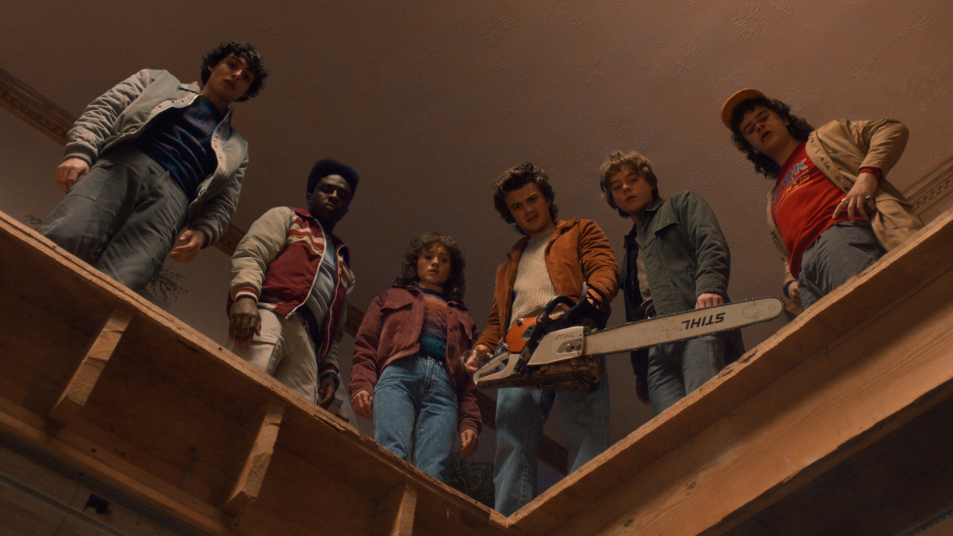 A group of teenagers (l-r: Finn Wolfhard as Mike Wheeler, Caleb McLaughlin as Lucas Sinclair, Natalia Dyer as Nancy Wheeler, Joe Keery as Steve Harrington, Charlie Heaton as Jonathan Byers, and Gaten Matarazzo as Dustin Henderson) look down from a wooden ledge, as one man holds a chainsaw, in STRANGER THINGS: SEASON 5.