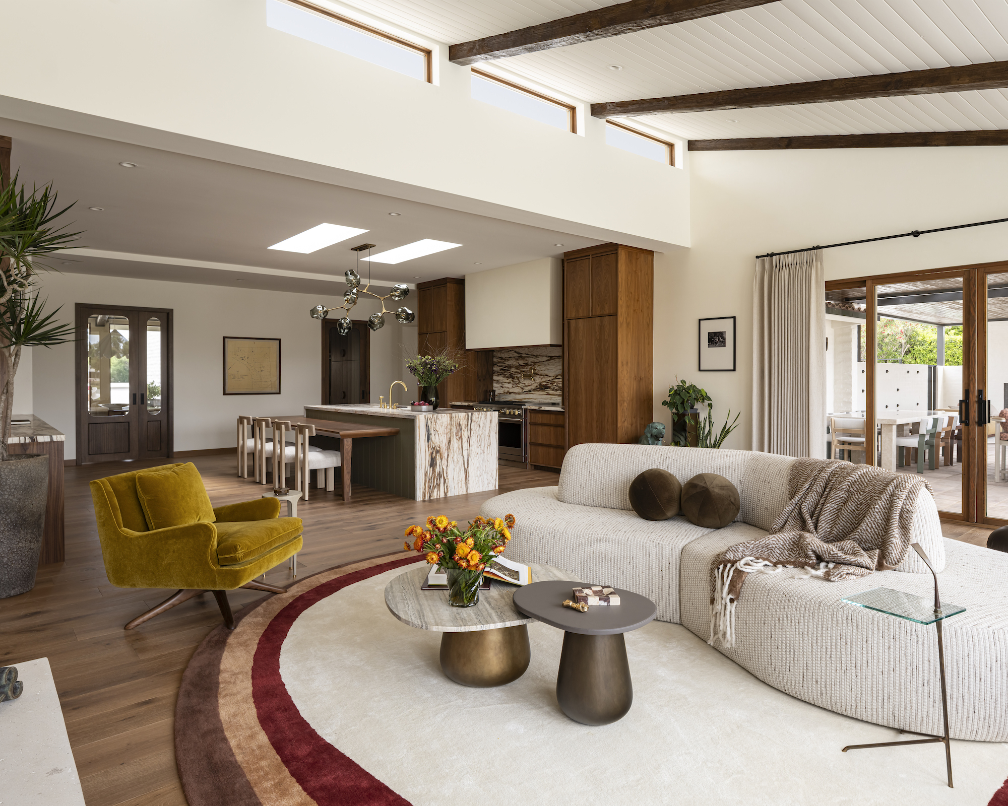 Living room with neutral curved sofa, mustard mid-century style armchair and dark wood kitchen beyond