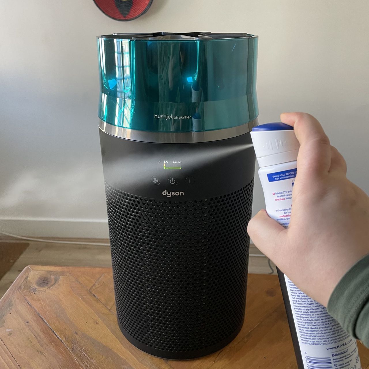 Hand spraying deodorant can in front of the Dyson Hushjet Air Purifier sitting on a wooden table