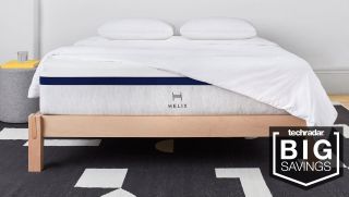 A Helix Sleep Midnight mattress shown on a light wooden bed frame and covered with white sheets and pillows