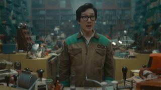 Screenshot from the Marvel T.V. show Loki. Ouroboros (Ke Huy Quan) standing behind a desk cluttered with rubber stamps. He has short dark hair, thick black rimmed glasses and is wearing an army green jumpsuit. The room behind him is full of bookshelves overflowing with all sorts of random trinkets.