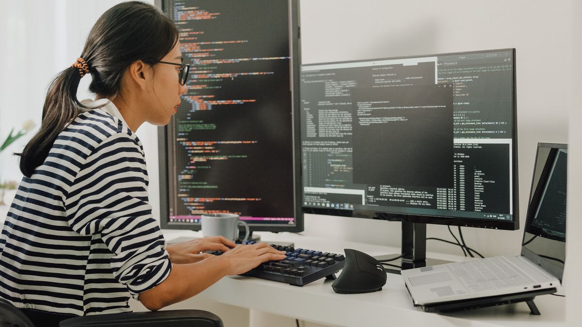 A woman software developer using computer to write code sitting at desk with multiple screens in different orientations