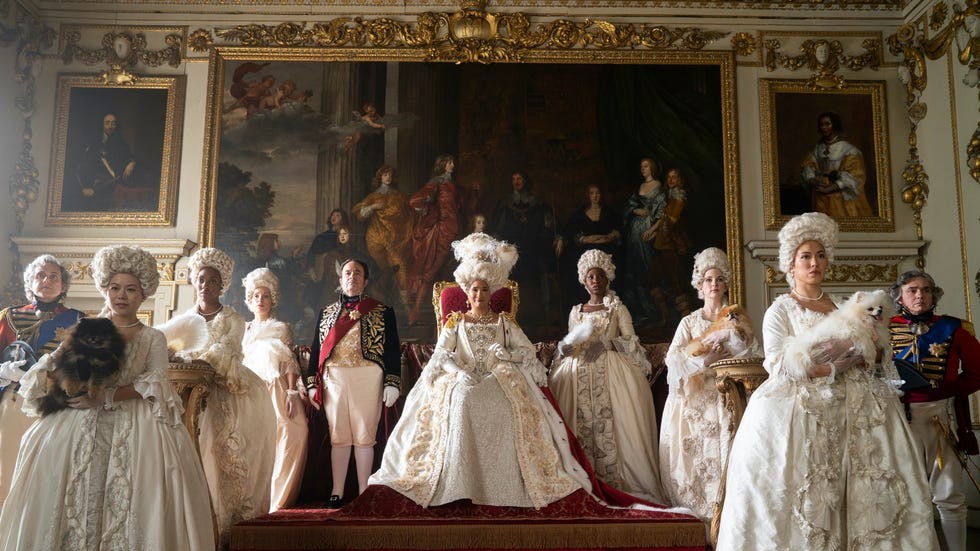 Scene from Bridgerton. A regal group in elaborate 18th-century-style gowns and wigs pose in a grand palace room with gold-framed portraits and large oil paintings, some holding fluffy lapdogs.