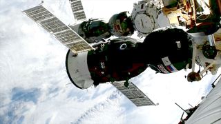 a newly-docked spacecraft is seen in the foreground, backdropped by a previous capsule at a space station in Earth orbit
