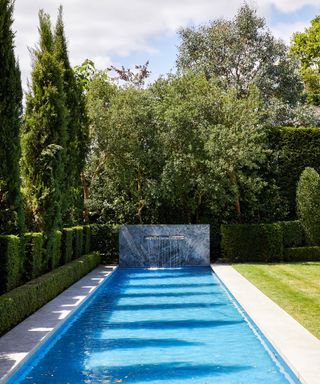 A swimming pool with a water feature at one end next to a hedge and lawn