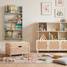 A playroom with an off-white bookcase with rattan inserts, a wall shelf and a rattan toy storage ox on a patterned rug in the middle