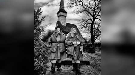 Black and white portrait of two elderly women in headscarves and heavy coats standing on stone steps before a tall wooden church spire, framed by bare tree branches