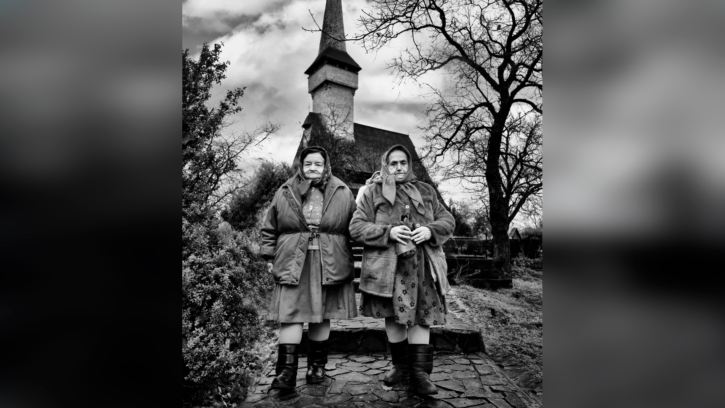 Black and white portrait of two elderly women in headscarves and heavy coats standing on stone steps before a tall wooden church spire, framed by bare tree branches