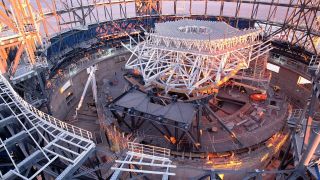 Looking down at the interior of a large circular structure under construction, filled with metal beams to makeup the skeleton of the structure.