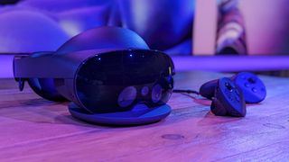 Et headset av typen Meta Quest Pro-headset ligger på et trebord ved siden av de tilhørende kontrollerne i et rom badet i blått lys med et skjær av rosa.