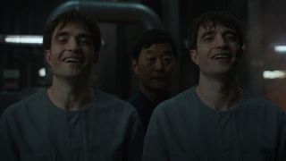 Scene from the movie Mickey 17. Two male clones wearing gray jumpsuits are standing next to each other whilst laughing.