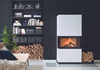 sleek, white and contemporary woodburning stove in a living space next to a storage unit with wood, papers and folders, against a dark black wall, on a white floor, with a bench to the right with cushions and a throw on it