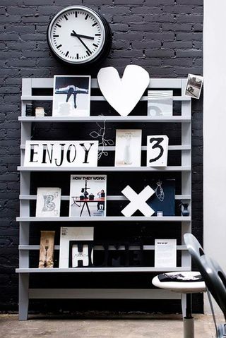 A free standing magazine rack with multiple shelves leant against a dark grey brick wall