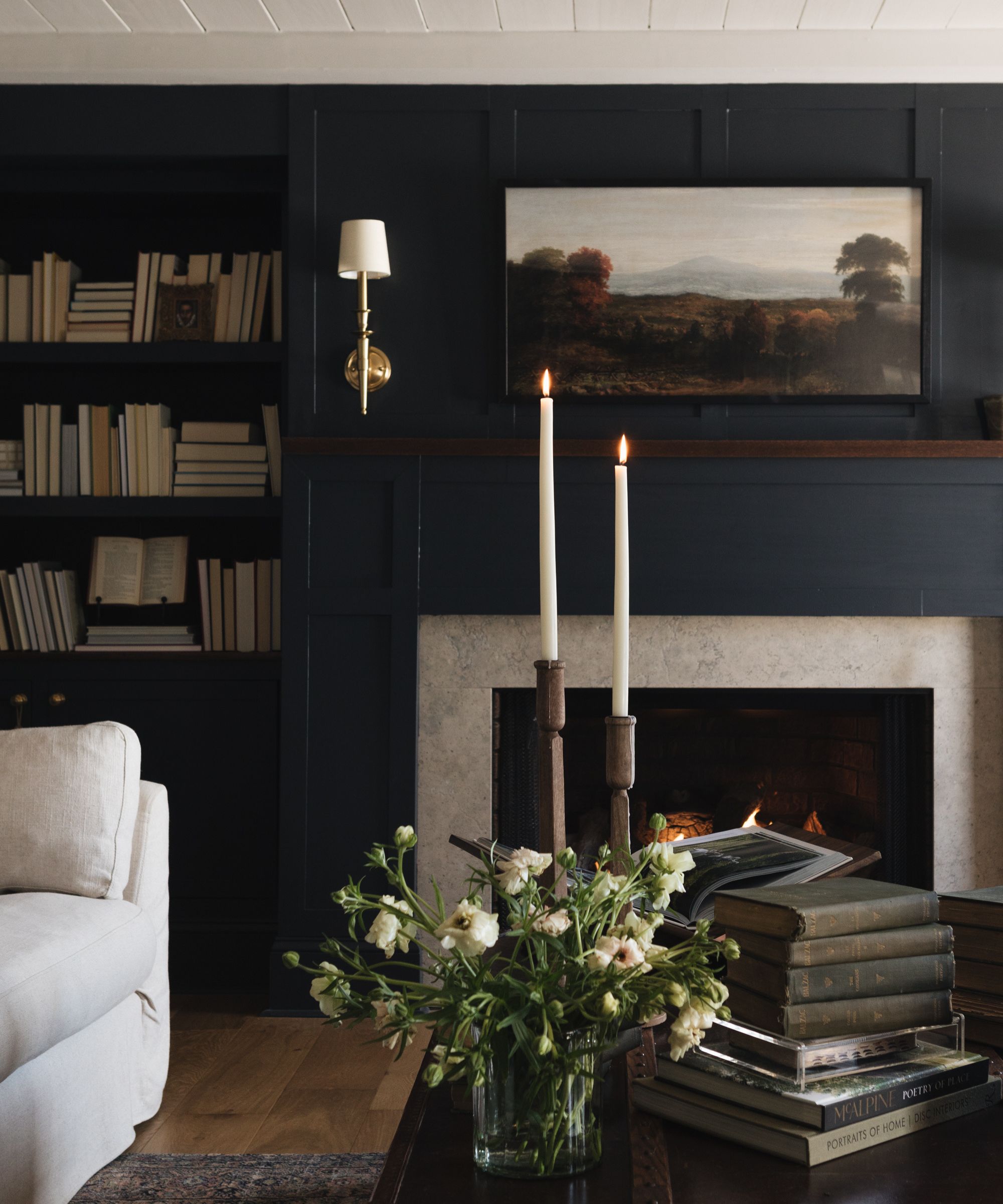 A cozy living room with dark blue-gray walls, a white ceiling, a fireplace with a landscape artwork above it, and bookshelves in the alcove.
