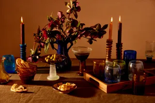 A vibrant table features multiple lit candleholders, flowers, a wooden tray, and colorful glasses.