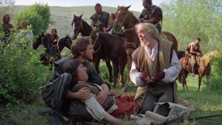 Still from the science fantasy movie Krull (1983). An injured man is lying down in a grassy field, being held and propped up by another man. Just in front of them is an older man who is getting bandages out of a bag. In the background is the rest of the group of adventurers, sitting on horseback (7 people visible).