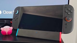 SIWIQU Switch 2 dock with handheld attached on black TV bench with screen in backdrop and shocked Kirby figure on left.