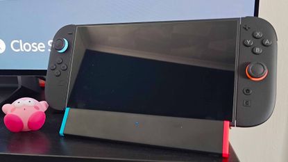 SIWIQU Switch 2 dock with handheld attached on black TV bench with screen in backdrop and shocked Kirby figure on left.