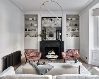 Living room with pale grey walls, bay window, built in storage either side of the chimney breast and fireplace. Grey rug and sofa and orange patterned armchairs.