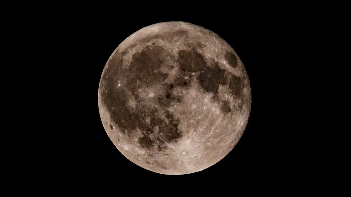 Una fotografía de la luna llena en un cielo negro que muestra mares lunares oscuros y cráteres de impacto brillantes.