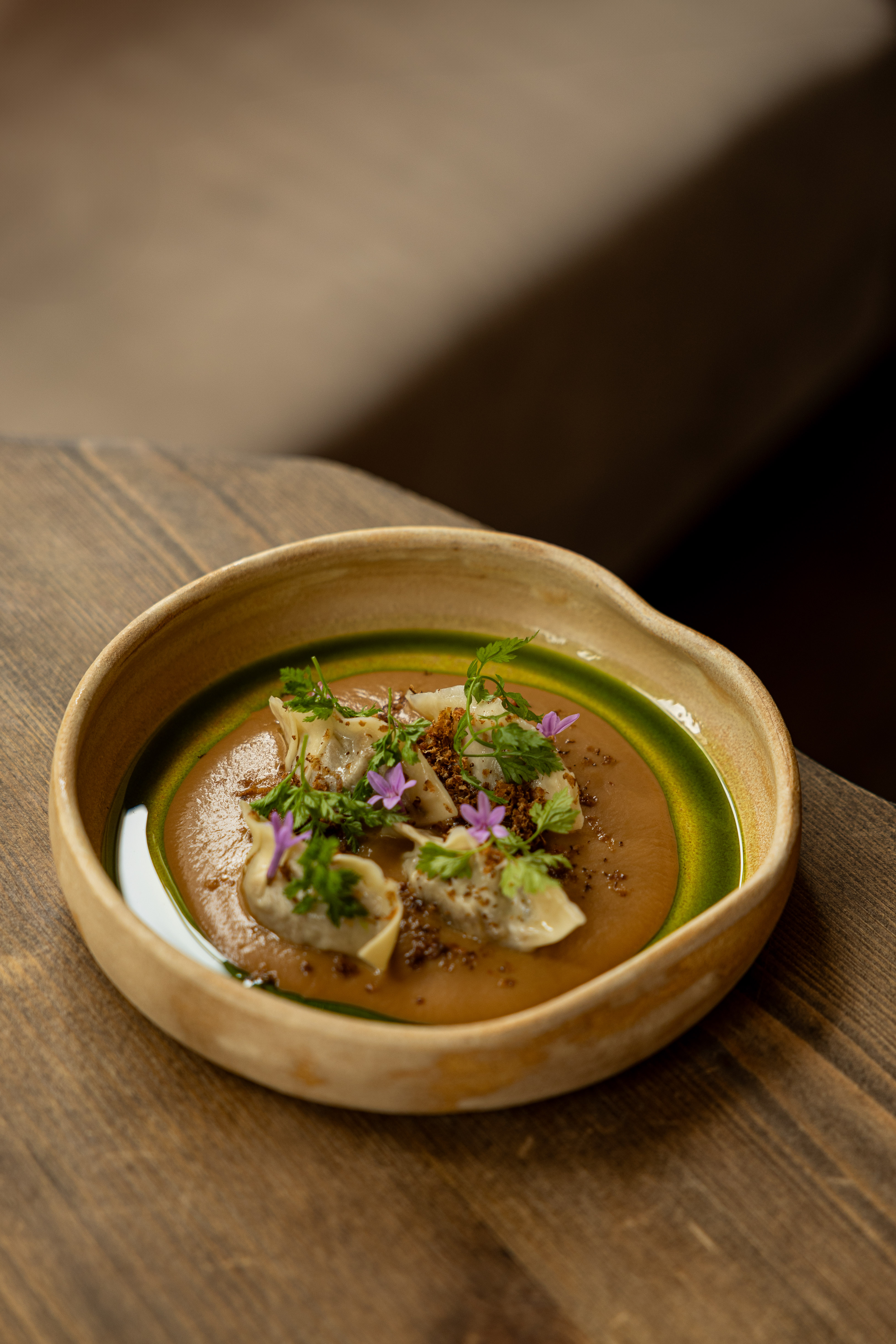 A beautifully plated dumpling dish, placed inside a neutral ceramic bowl, boasts a basil-infused oil topping, flowers, and fresh herbs.