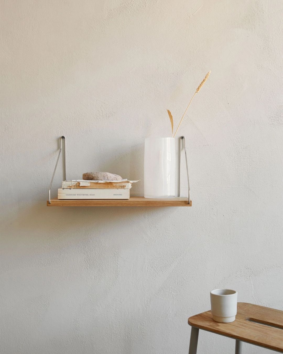 A wooden shelf with chrome arms, hung up on a plain beige wall and topped by books, some bread, and a vase, next to which sits a wooden bench topped by a white coffee mug.