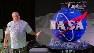 a man in a white t-shirt stands next to a large spherical cake that looks like nasa's famous "meatball" logo