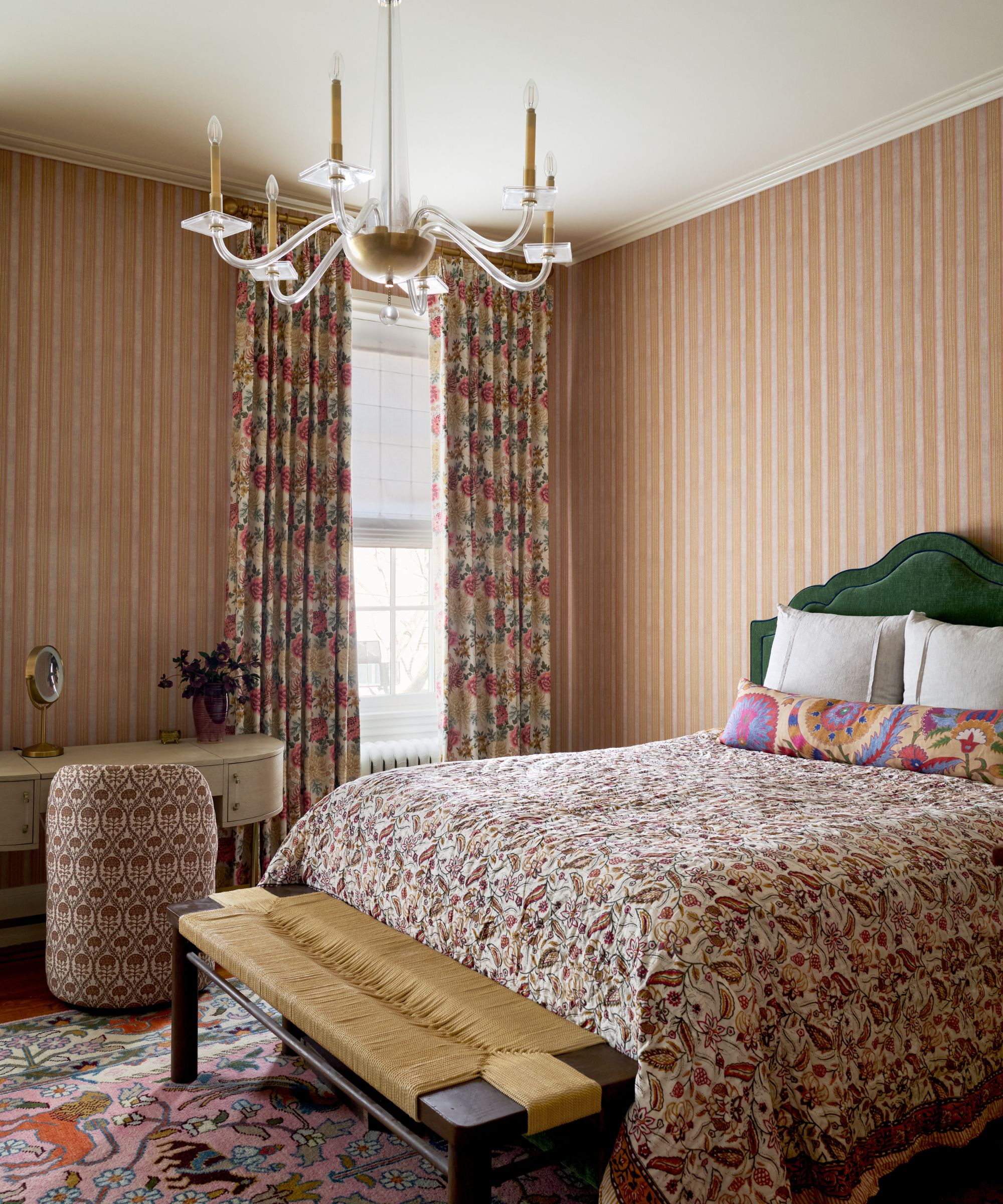 Maximalist bedroom with pink striped wallpaper, a green velvet headboard, a colorful paisley quilt, and a glass chandelier.