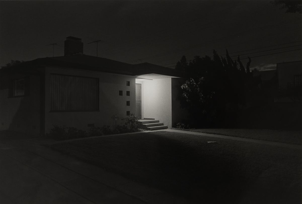 A black and white photograph of a house at night, its entryway lit by a single bright light against dark surroundings and trees.