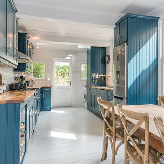 Wooden kitchen table and chairs on wooden floor in in blue kitchen with white door with glass with a garden view