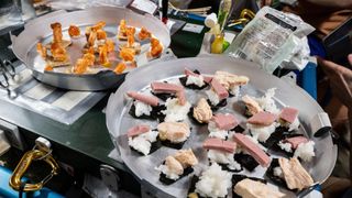 A metal plate full of sushi with seaweed, rice, and spam sit on a table next to another plate with shrimp tails on crackers served on it