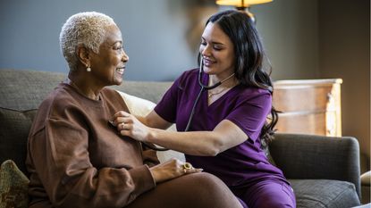 A smiling woman sitting on a couch is checked over by a smiling nurse.