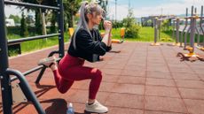 woman wearing red leggings and black top doing bulgarian split squat in an outdoor park gym